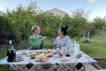 A blonde haired women in a green shirt sits on the left, touching her glass with a dark haired woman in a blue shirt on the right. They are sitting at an outdoor table surrounded by trees, with a table cloth and various food goods on the table in front of them.